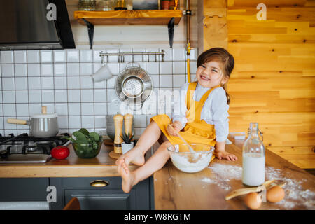 A little cute girl pretending to bake a cake in the kitchen. Sitting on the table beside flour, milk and a bowl with dough. Stock Photo