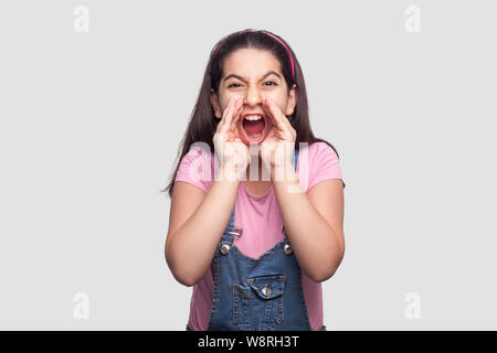Portrait of screaming brunette young girl in casual style, pink t-shirt and blue denim overalls standing looking with hand near mouth and shouting. in Stock Photo