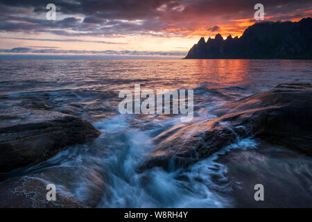 Waves crushing over mighty rocks with impressive oksen Mountains in background at a colorful midnight sunset, Tungeneset, Devils Jaw, Senja, Norway Stock Photo