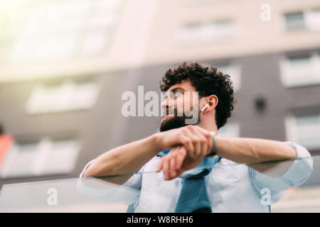 Successful man in a blue suit stands leaning on the railing next to modern glass buildings resting outdoors. Wide shot. Stock Photo