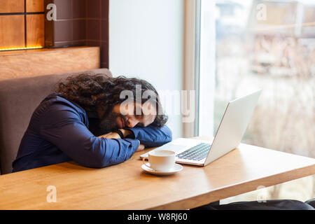 Tired handsome young adult man freelancer in casual style sitting in cafe with laptop, sleeping on the table after hard overtime work, closed eyes, bu Stock Photo