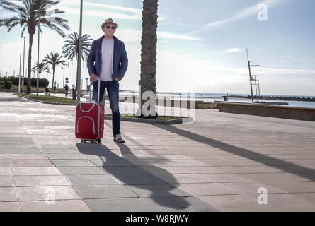 Attractive serious man stands with a red suitcase on the embankment Stock Photo