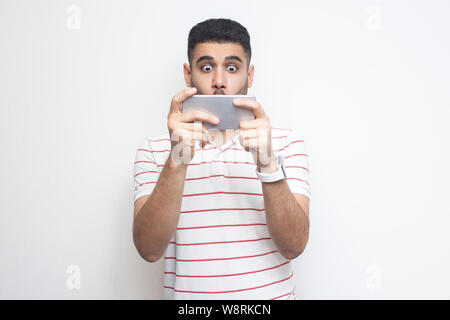 Exited gamer young adult man wearing in white T-shirt standing, using smartphone and playing mobile game with upset face and big eyes, covering mouth. Stock Photo