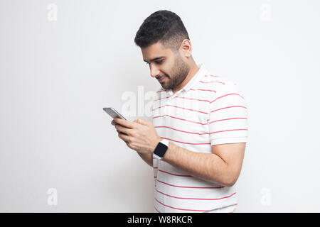 Side view portrait of happy handsome blogger young adult man wearing T-shirt standing, using smartphone and texting message with smile. Indoor, isolat Stock Photo