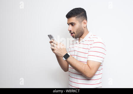 Side view portrait of shocked handsome blogger young adult man wearing in white T-shirt standing, using smartphone and watching video with amazed face Stock Photo