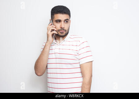 Portrait of serious handsome young adult man wearing T-shirt standing, using smartphone, making call and talking with friend. Indoor, isolated, studio Stock Photo
