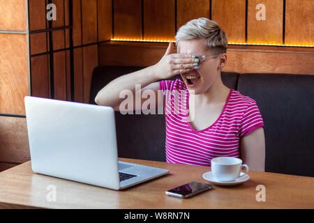 Portrait of emotional scared happy young businesswoman in pink t-shirt is sitting in cafe and covering the eyes to herself with hand and laughing caus Stock Photo