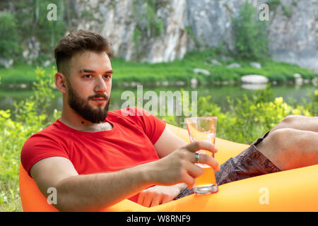 Leisure concept, camping, summer vacation. Young bearded caucasian man with glasses of beer laying at orange sofa. Relaxing and enjoying life on air b Stock Photo