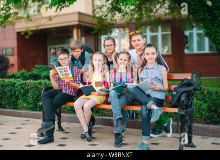 Happy Schoolmates Portrait. Schoolmates seating with books in a wooden bench in a city park and studying on sunny day. Stock Photo