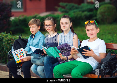Happy Schoolmates Portrait. Schoolmates seating with books in a wooden bench in a city park and studying on sunny day. Stock Photo