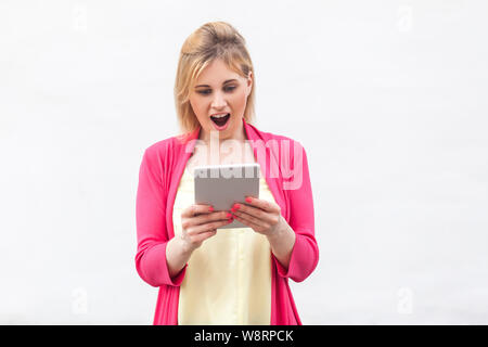 No way! Portrait of beautiful unbelievable young woman in pink blouse standing and watching suprised video on her tablet with shocked face. Indoor, is Stock Photo