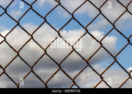 Old rusty mesh netting on a blue sky with clouds, background wallpaper texture. Rust on the metal fence of the danger zone Stock Photo