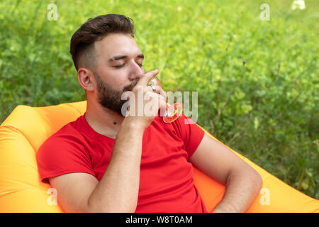 A young man with a beard lies on an orange Lamzac inflatable sofa and drinks a white alcoholic drink from a glass. Enjoys outdoor recreation Stock Photo