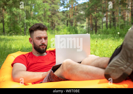 A bearded man, a businessman sitting with a laptop on an inflatable sofa on the grass against the background of the forest. Freelance Concept, Remote Stock Photo