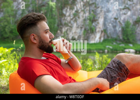 A young man with a beard lies on an orange inflatable sofa Lamzak and drinks beer from a glass on the shore. at the picnic enjoys relaxation Stock Photo