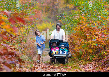 Family hiking with stroller in autumn park. Active father, baby and toddler in twin double pushchair. Fit healthy dad walking with jogger pram and kid Stock Photo