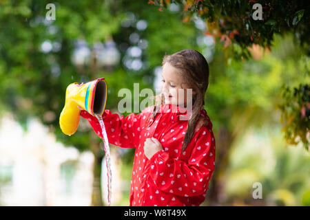 Kid with rubber boots playing in the rain in autumn park. Child in muddy puddle on rainy fall day. Rain boot full of water. Little girl in red jacket Stock Photo