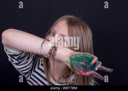Beautiful blond young girl with freckles and hands painted in colorful paints indoors on black background, closeup portrait