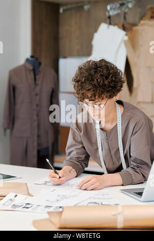 Young brunette female bending over desk while making fashion sketches Stock Photo