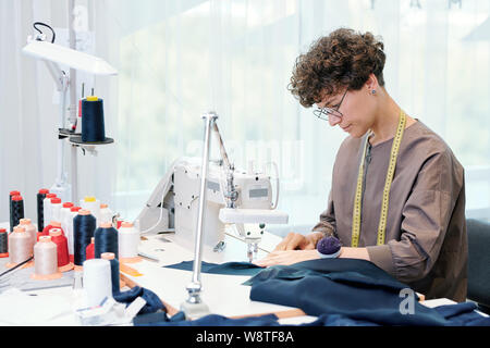 Pretty young seamstress sitting by sewing machine and processing blue textile Stock Photo