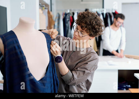 Young serious seamstress stitching unfinished blue dress on mannequin Stock Photo