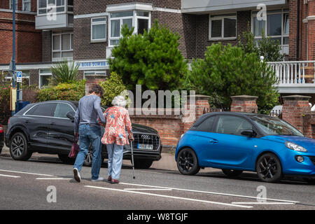 A man helping an elderly lady with a walking stick or cane to cross a busy road, Older/disabled person with carer Stock Photo