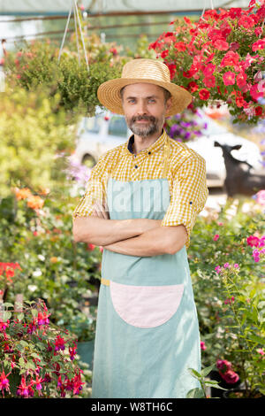 Mature male farmer or gardener in apron and hat standing in greenhouse Stock Photo