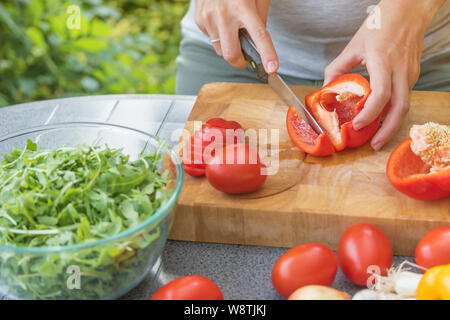 Closeup view of female hands slicing a red pepper on a wooden board. Various types of vegetables are lying on the table. Stock Photo