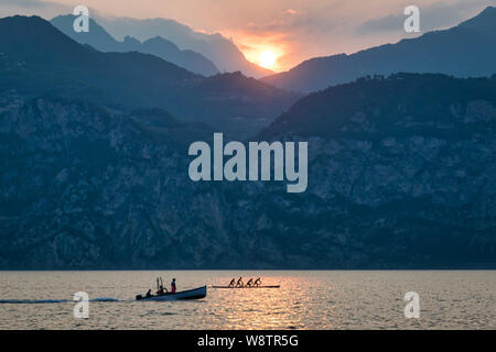 This is a sunset picture of Lake Garda.  Int the forground there is a fishing boat and a rowing team. In the background is the sun setting behind the Stock Photo