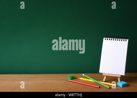Back to school concept, open notebook on miniature easel and several colored pencils on wooden surface on the backdrop of a clean green chalk board, s Stock Photo