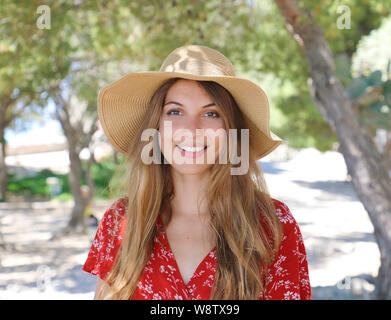 Portrait of a beautiful smiling girl wearing red dress and hat looking at camera when walking under olive trees Stock Photo