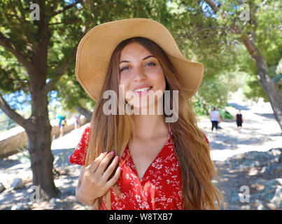 Beautiful smiling girl wearing red dress and hat looking at camera when walking under olive trees Stock Photo