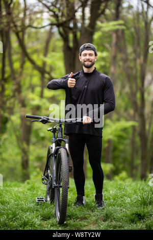 Happy man shows thumb up, riding on bicycle in pine forest, sport leisure. Cyclist walk in park, active healthy lifestyle Stock Photo