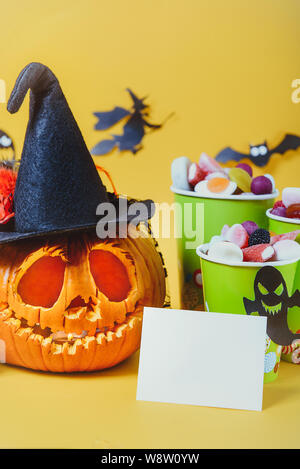 Carved pumpkin in hat, blank card and cups with sweets, candies decorated with silhouettes of bats, witch, ghosts on yellow background. Spooky holiday Stock Photo