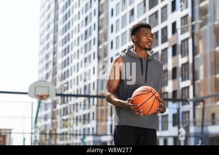 Waist up portrait of masculine African man holding basketball ball in court outdoors, copy space Stock Photo