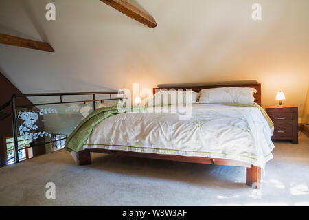 King size bed on teak wood frame with end tables, black wrought iron and tempered glass balustrade, old wooden structural beams in master bedroom Stock Photo