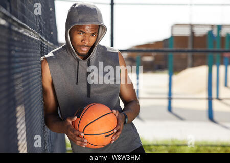 Waist up portrait of contemporary African-American man holding basketball ball looking at camera while posing in sports court outdoors, copy space Stock Photo