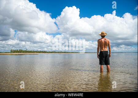 Tourist wearing a straw sun hat standing in calm shallow waters on the shore of a sunny palm-lined tropical island beach Stock Photo