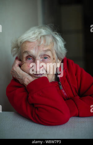 Old lady woman in her house, a portrait sitting at a table in a red jacket. Stock Photo
