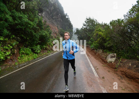 Male runner jogging and running on road in rain in jacket and long tights. Fit fitness model man working out living healthy lifestyle training for marathon. Young caucasian model in his 20s. Stock Photo