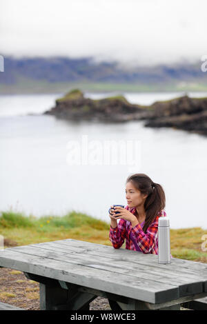 Camping woman sitting at table drinking coffee from thermos bottle flask by lake on Iceland. Camper girl relaxing thinking pensive taking break on road trip in beautiful Icelandic nature. Stock Photo