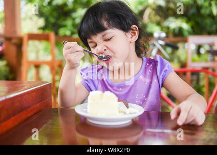 Cute little girl eating cake / Asian child happy and holding a spoon into the mouth with cake on dining table , selective focus Stock Photo