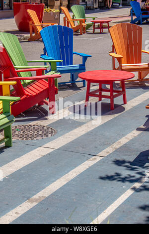 Brightly-coloured wooden chairs surround round tables on a street in a downtown marketplace on a sunny day. Stock Photo