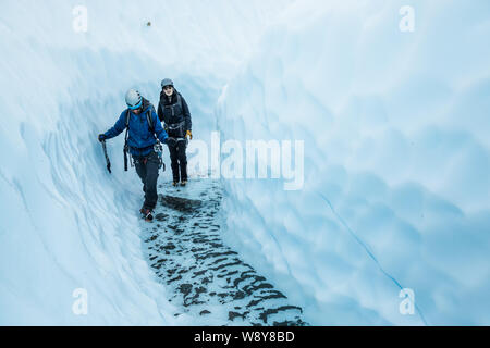 Young man and woman walking through a canyon of glacier ice on the Matanuska Glacier. In the bottom of the canyon is clear, cold water deep enough to Stock Photo