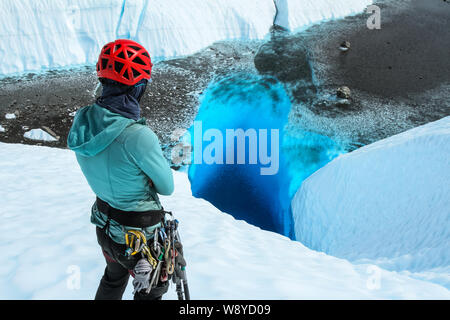 A young woman stands with arms crossed staring down a deep blue hole filled with water on the Matanuska Glacier in Alaska. Stock Photo