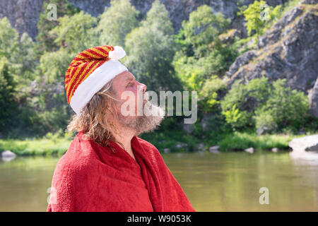 An older or charismatic elderly man with a beard in a Santa Claus hat and red coat looks up. Against the background of a summer landscape, river, moun Stock Photo