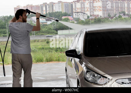 A young man in a gray washes his car at a car wash. City service for self-cleaning passenger cars sedan. A young man with a beard pours water on a car Stock Photo