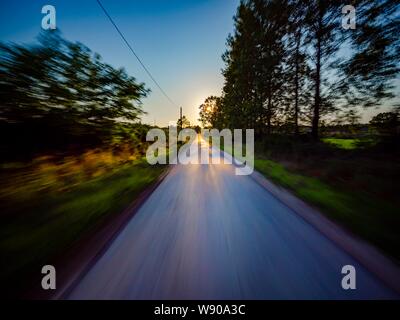 Sunset countryside drive narrow road passing through Green forest natural environment speeding up towards the setting sun Stock Photo