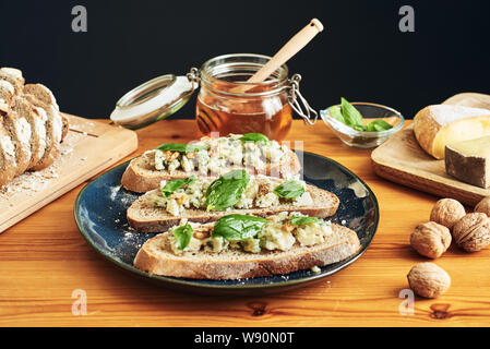 Close up on a plate of toasts with roquefort cheese, walnuts, honey and basil. Some cheese, nuts and honey in the background Stock Photo