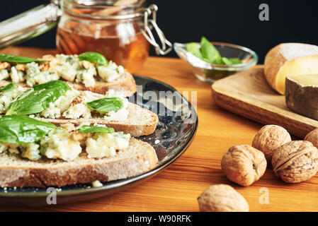 Close up on a plate of toasts with roquefort cheese, walnuts, honey and basil. Some cheese, nuts and honey in the background Stock Photo
